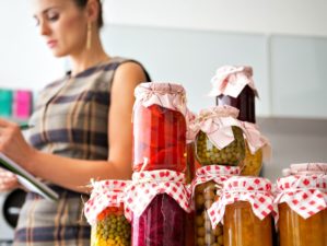 Featured | Housewife writing in notepad with jars of homemade fruits jam and pickled vegetables | Items to Kick-Start Your Food Storage Plan