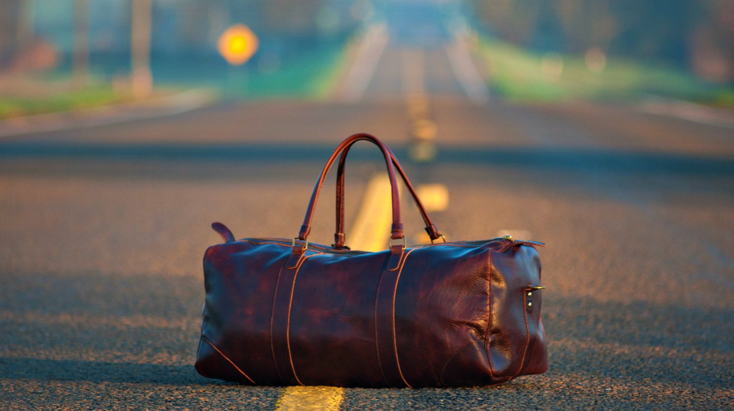 Feature | A brown leather duffel bag in middle on gray asphalt road | Preparedness Tips: Prepping For Travel