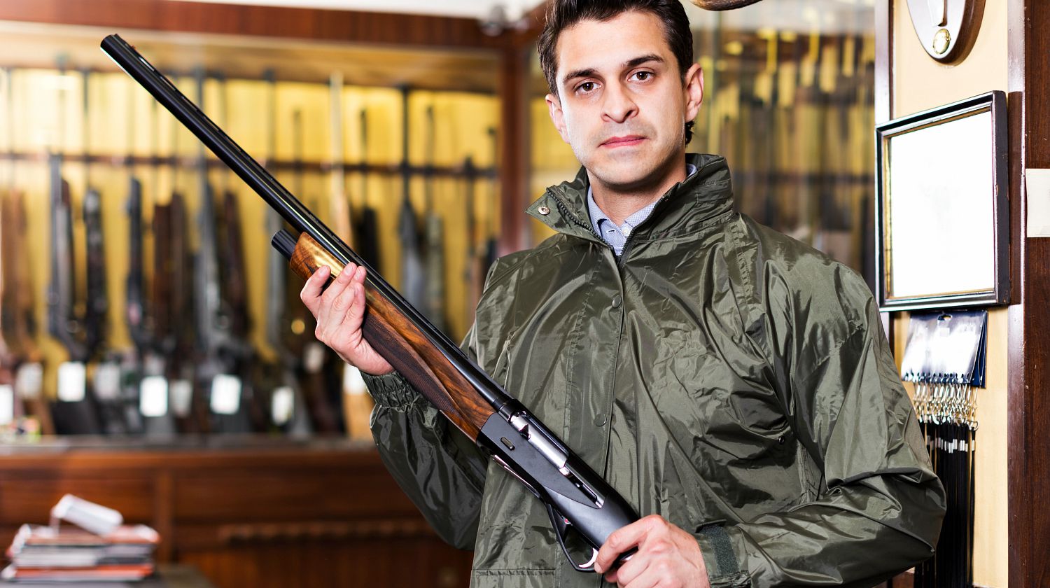 Portrait of positive man in hunting raincoat in gun shop carefully picking out rifle | Places To Buy Guns Online, And A Few Tips To Help | Featured