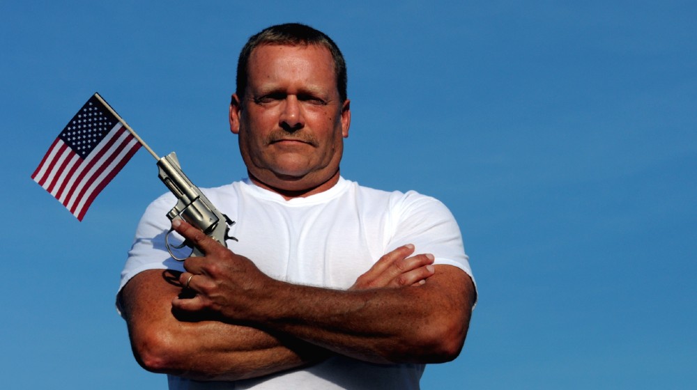 Muscular White Man Holding An American Flag And Gun, symbolizing our constitutional right to own and bear arms-Gun Owner-ss-featured